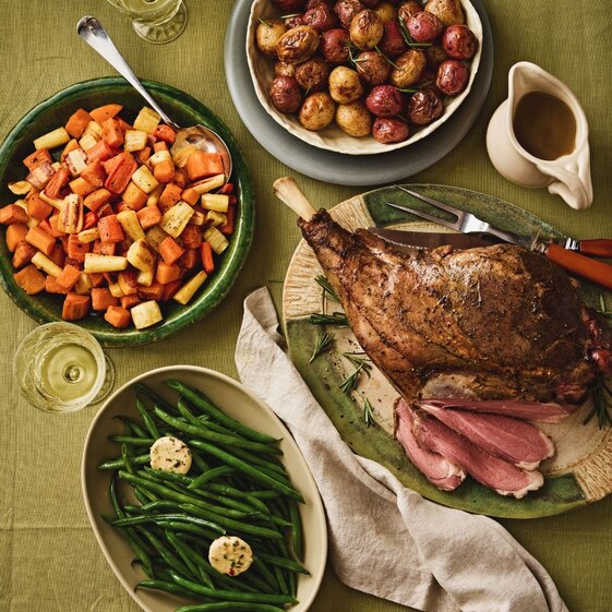 Roast lamb dinner with roasted root vegetables, baby potatoes, green beans, and gravy on a family meal table.